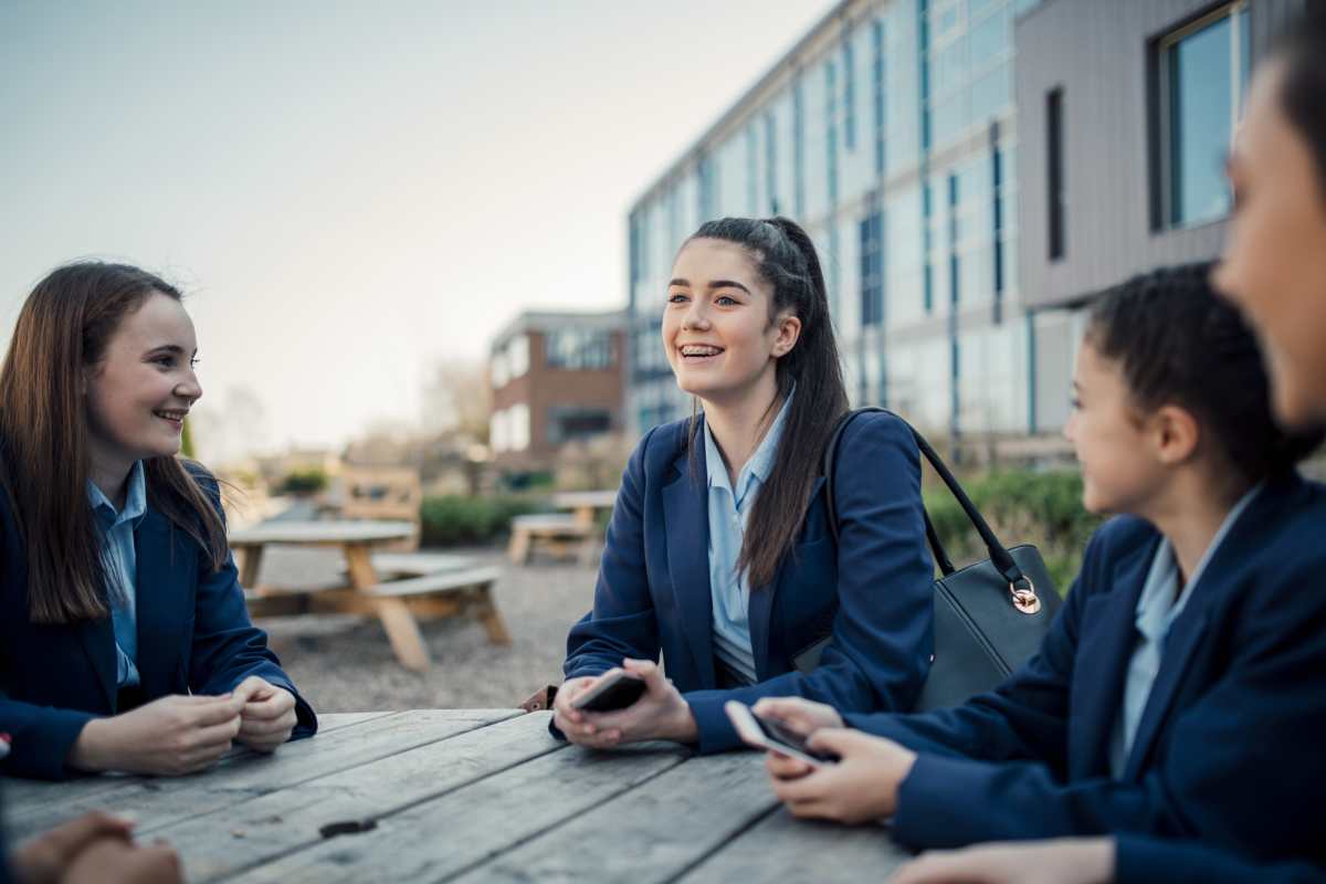 A group of schoolgirls sit talking at a table outside of school

Cover Image Source: Getty Images | Photo by SolStock